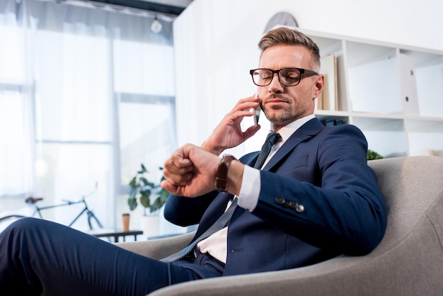 Empresario con gafas sentado en un sillón y mirando el reloj mientras habla por teléfono inteligente