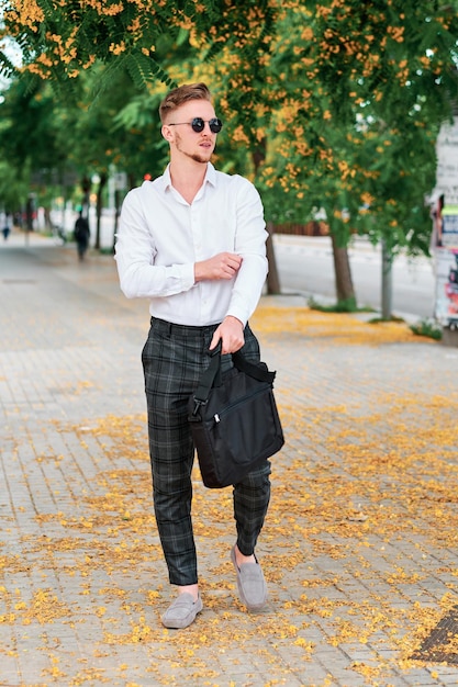 Empresario con gafas de sol y sosteniendo una bolsa mientras camina al aire libre en la calle