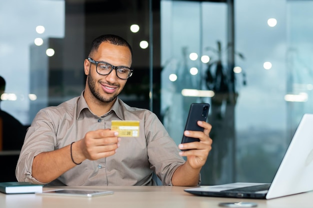 Empresário hispânico bem-sucedido dentro do escritório homem de camisa sorrindo e feliz segurando o crédito bancário