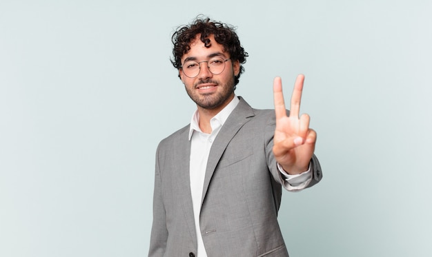 Foto empresário hispânico sorrindo e parecendo feliz, despreocupado e positivo, gesticulando vitória ou paz com uma mão