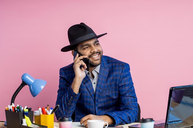 Empresario indio con estilo alegre vistiendo ropa formal, sombrero negro, hablando por teléfono móvil, sonriendo ampliamente, mirando a un lado, sentado en el escritorio en la oficina contra la pared rosa. Trabajo, concepto de negocio.