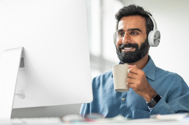 Empresario indio tomando café en el lugar de trabajo usando auriculares mientras trabaja en una computadora en