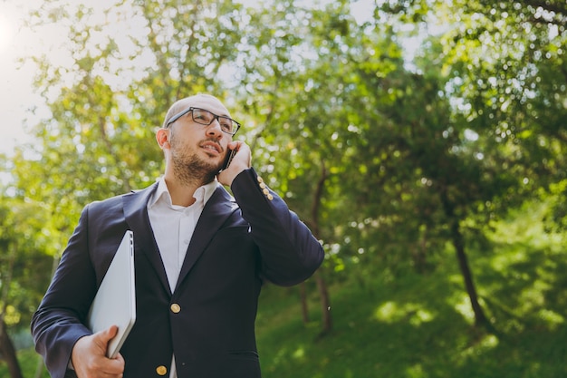 Empresário inteligente bem sucedido na camisa branca, terno clássico, óculos. Homem ficar com o computador laptop pc, falar no celular no parque da cidade ao ar livre no fundo da natureza. Escritório móvel, conceito de negócio.