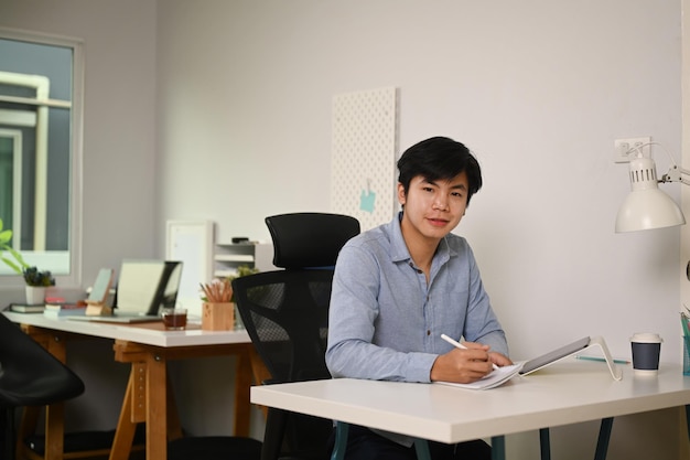 Empresário inteligente segurando tablet digital e sorrindo para a câmera enquanto está sentado no escritório em casa