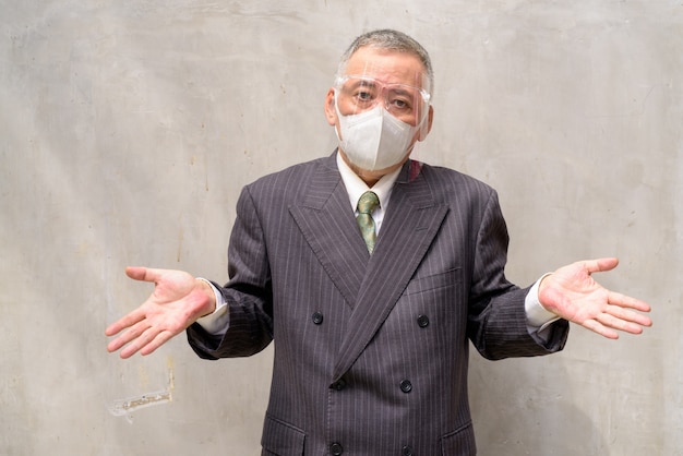 Empresário japonês maduro com máscara e escudo do rosto encolher os ombros os ombros
