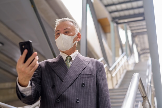 Empresario japonés maduro con máscara usando el teléfono mientras baja las escaleras en la ciudad