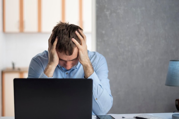 Empresário jovem trabalha com laptop e mantém as mãos na cabeça cansadamente. Problemas no trabalho. Esgotamento profissional.