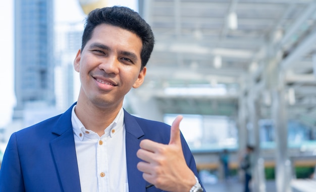 Empresario latino muestra gesto de la mano pulgar hacia arriba (buena señal) en el fondo del edificio de la ciudad exterior