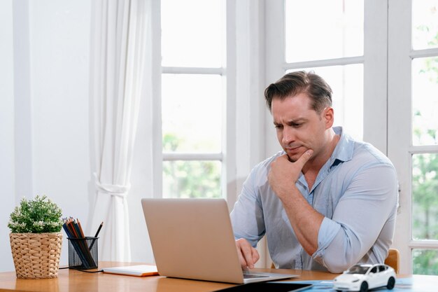 Empresário luta para resolver problemas de trabalho em casa usando laptop sentado em sua mesa no escritório em casa com expressão estressada e frustrada tentando descobrir uma solução para o problema Synchronos