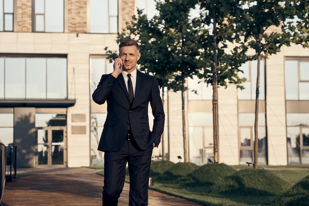 Empresário maduro confiante falando no celular enquanto caminha perto de um prédio de escritórios ao ar livre