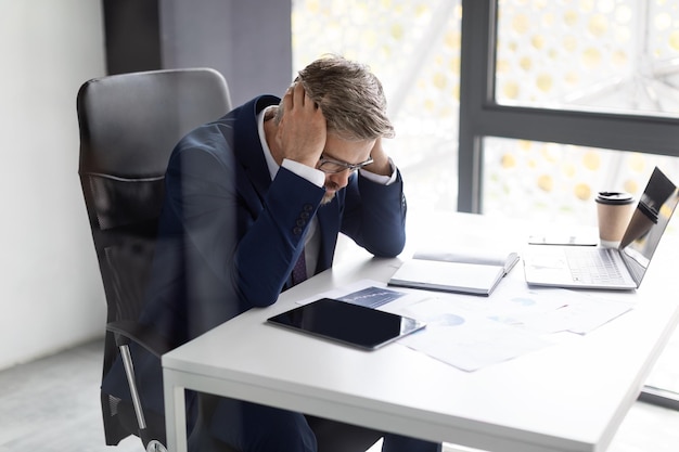 Empresario maduro estresado en traje tocando la cabeza mientras está sentado en el lugar de trabajo