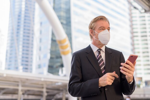 Empresario maduro con máscara pensando mientras usa el teléfono en el puente skywalk