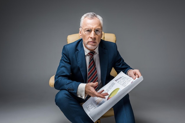 Foto empresario maduro en traje y anteojos sosteniendo gráficos y gráficos mientras está sentado en un sillón en gris
