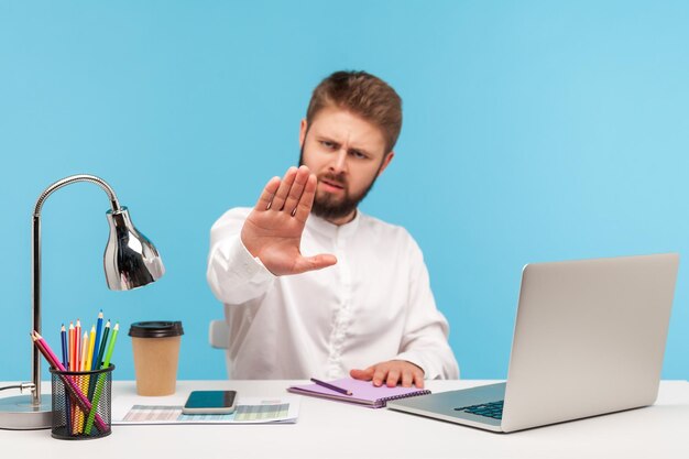 Empresário mandão estrito com barba sentado no local de trabalho do escritório com laptop mostrando parada, aviso com gesto de mão, acesso negado, proibição. Tiro de estúdio interior isolado em fundo azul