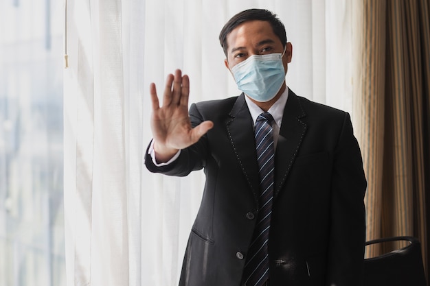 Empresario de máscara médica con traje negro y corbata de pie con gesto de parada con la mano