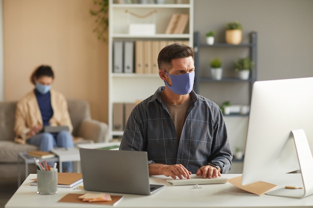 Empresario de máscara protectora trabajando en equipo en su mesa con su colega en la pared de la oficina
