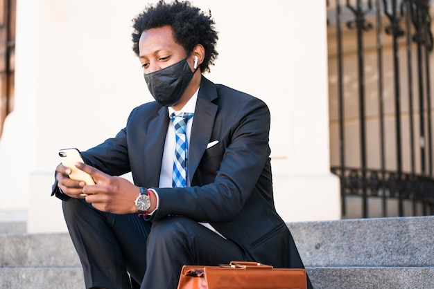 Empresario con mascarilla y usando su teléfono móvil mientras está sentado en las escaleras al aire libre