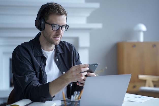 Empresario masculino pensativo en auriculares sentado en el escritorio en la oficina en casa y usando teléfono móvil