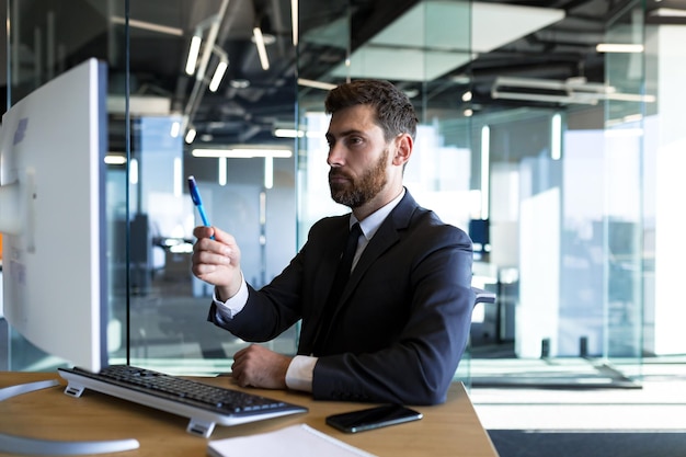 Empresario masculino serio y reflexivo que trabaja en la oficina en la computadora con un traje de negocios