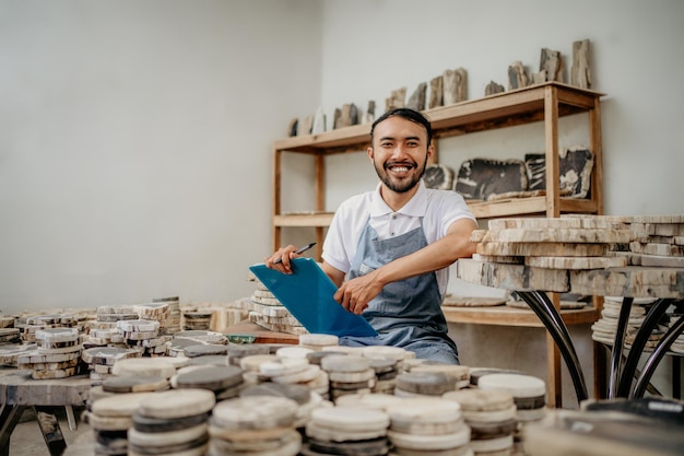 Empresario masculino sonriendo a la cámara sentado con un portapapeles y un bolígrafo en un almacén de artesanía de piedra