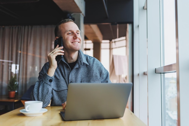 Empresario masculino sonriente con ropa informal sentado en la mesa con una laptop y hablando por teléfono inteligente en un espacio de trabajo moderno con una ventana grande durante el día