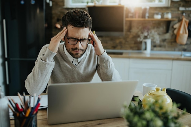 Empresario masculino sosteniendo su cabeza con dolor mientras trabaja en una computadora portátil en casa