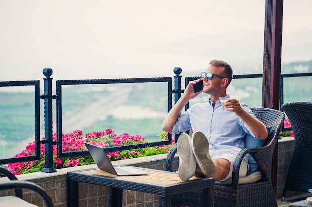 Empresário masculino trabalhando em um laptop de férias com uma bela vista panorâmica Gerente bem sucedido bebendo café e falando ao telefone com café enquanto viaja