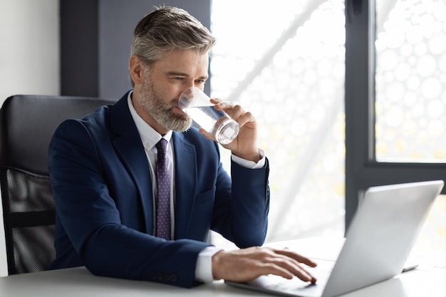 Empresario de mediana edad bebiendo agua mientras trabaja con un portátil en la oficina