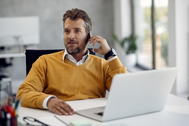 Empresario de mediana edad hablando por teléfono mientras trabaja en una laptop en la oficina