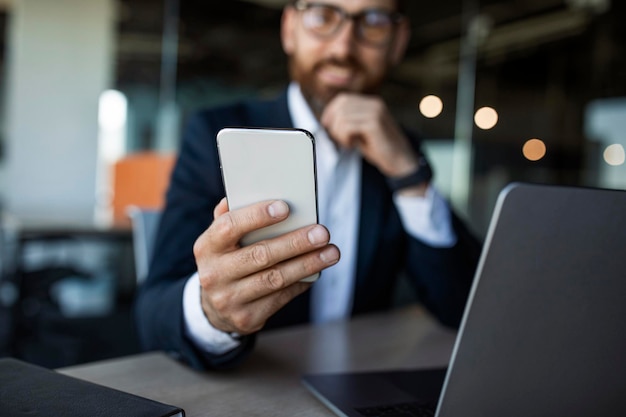 Foto empresario de mediana edad sentado en el escritorio y sosteniendo el teléfono celular en la mano enviando mensajes de texto con el enfoque selectivo de los clientes