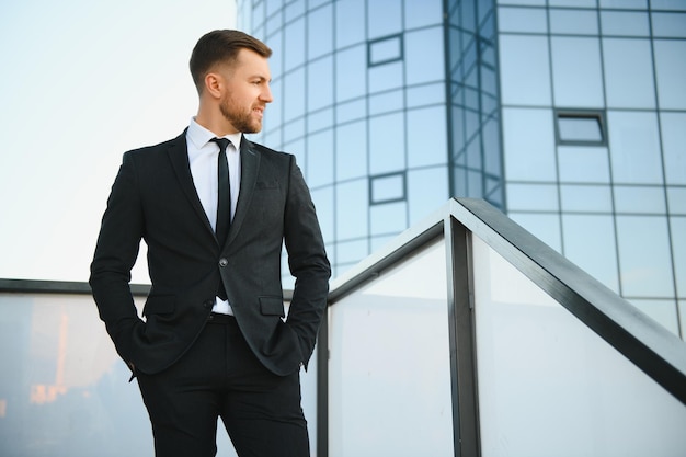 Empresário moderno Jovem confiante de terno completo e olhando para longe enquanto está ao ar livre com a paisagem urbana ao fundo
