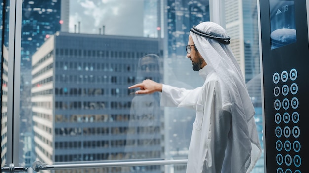 Foto empresário muçulmano de sucesso em kandura branco tradicional montando um elevador de vidro para o escritório em um centro de negócios moderno homem árabe interagindo com uma maquete de tela holográfica no conceito do elevador