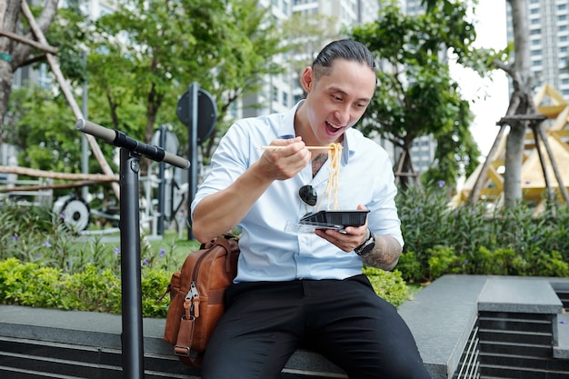 Empresario multiétnico positivo sentado en un banco cerca del edificio de oficinas y comiendo fideos para llevar