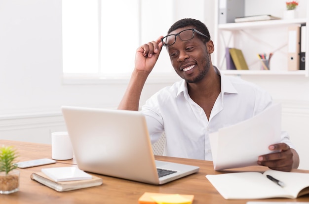 Empresário negro no escritório, usando óculos, trabalhando com laptop e papéis com prazer