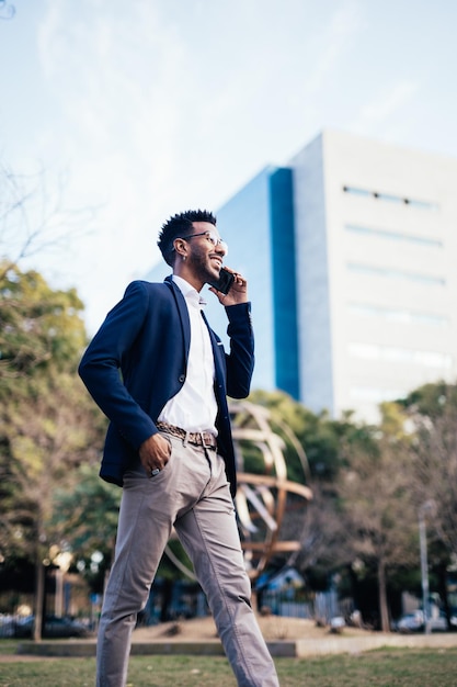 Empresário negro sorridente andando por um parque enquanto usa seu telefone fazendo negócios