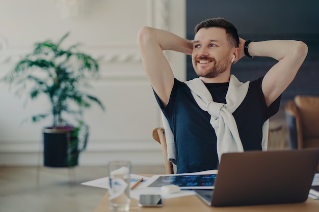 Empresario o autónomo despreocupado listo para trabajar mientras está sentado en su acogedor lugar de trabajo en casa con una computadora portátil y documentación del proyecto. Hombre manteniendo las manos detrás de la cabeza y mirando a un lado