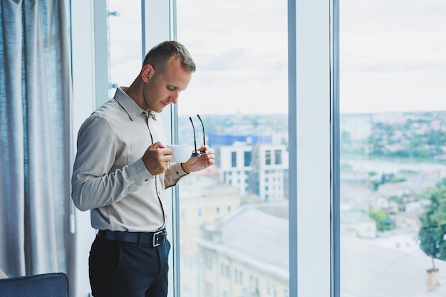 Empresario de oficina moderno o gerente bebiendo café junto a la ventana sonriendo Retrato de un exitoso programador de ingenieros de TI con gafas trabajando en la oficina