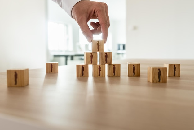 Empresário, organizando cubos de madeira com silhueta de pessoas em forma de pirâmide com outras pessoas espalhadas na mesa de escritório Conceitual de recursos humanos de organização empresarial e trabalho em equipe