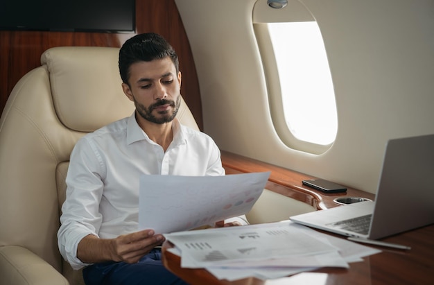 Empresário pensativo do Oriente Médio segurando um relatório financeiro, trabalhando com documentos dentro de um avião