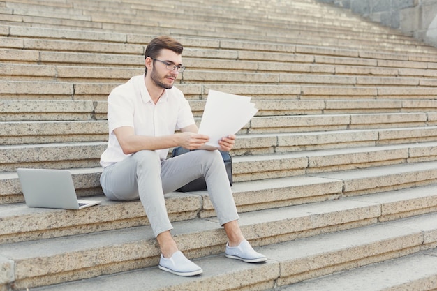 Empresário pensativo em óculos está lendo documentos de contrato enquanto está sentado entre o laptop e a maleta nas escadas de pedra, copie o espaço. Conceito de tecnologia e negócios