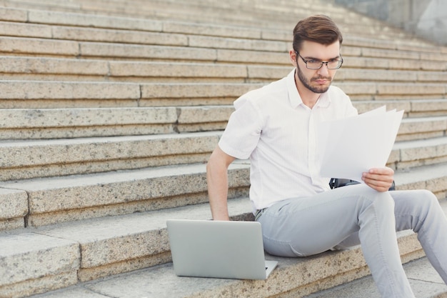 Empresário pensativo em óculos está lendo documentos do contrato e digitando em seu computador portátil enquanto está sentado nas escadas de pedra, closeup, copie o espaço. Conceito de tecnologia e negócios