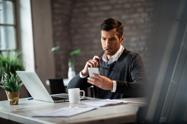 Empresario pensativo leyendo mensajes de texto en el teléfono móvil mientras trabaja en la oficina