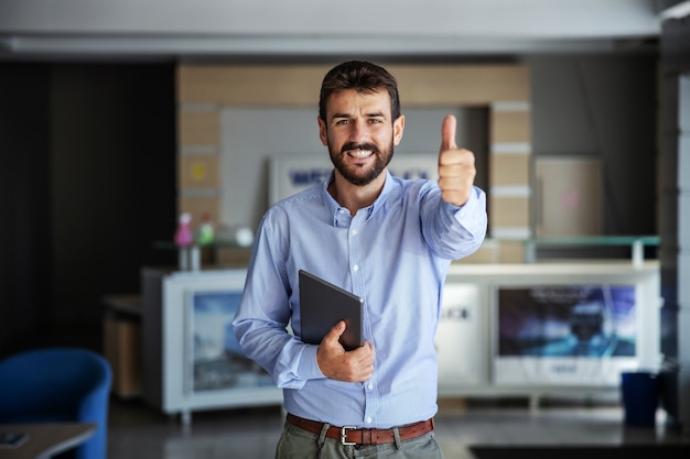 Empresario de pie en el vestíbulo de la empresa de exportación, sosteniendo la tableta y mostrando los pulgares para arriba.