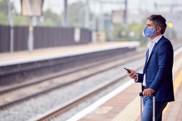 Empresario en la plataforma ferroviaria con teléfono móvil con máscara facial PPE durante la pandemia de salud