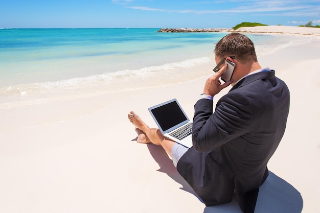 Empresario en la playa trabajando con computadora y hablando por teléfono