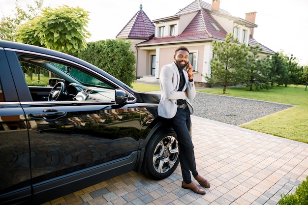 Empresário preto bonito de terno falando pelo smartphone perto de um carro preto, ao ar livre, construindo