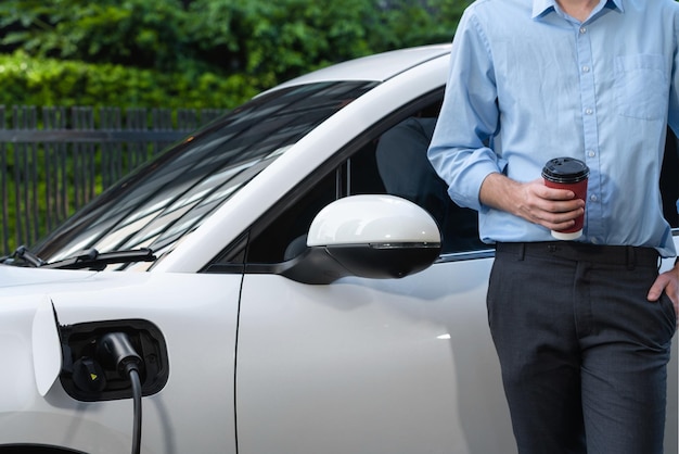 Empresário progressivo com café e carro EV na estação de carregamento