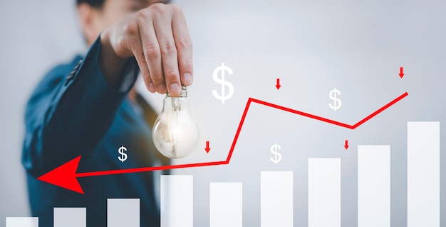 Foto empresario que sostiene una bombilla con pérdidas en el concepto de gráfico bursátil, gráfico de mercado y flechas rojas caen al suelo, lo que indica la recesión económica que ocurrirá en 2021