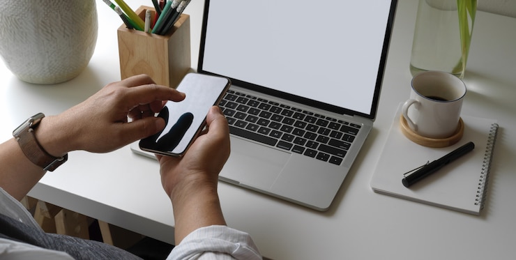 Foto empresario que usa un teléfono inteligente mientras está sentado en un simple escritorio de oficina con computadora portátil, agenda, papelería y decoraciones
