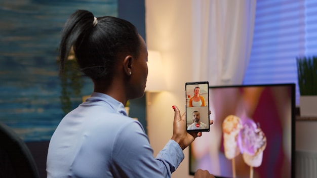 Empresario remoto de mujer negra con videoconferencia con un amigo usando un teléfono inteligente sentado en la sala de estar a altas horas de la noche. Empleado ocupado utilizando tecnología inalámbrica de red de tecnología moderna haciendo horas extraordinarias para el trabajo